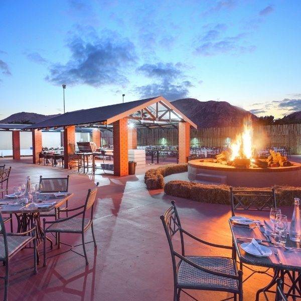 Photo of ANEW's Hunters Rest Resort in Rustenburg, with tables and chairs set on an open braai area with a communal fire pit behind them, set against the backdrop of the sky reaching dusk.